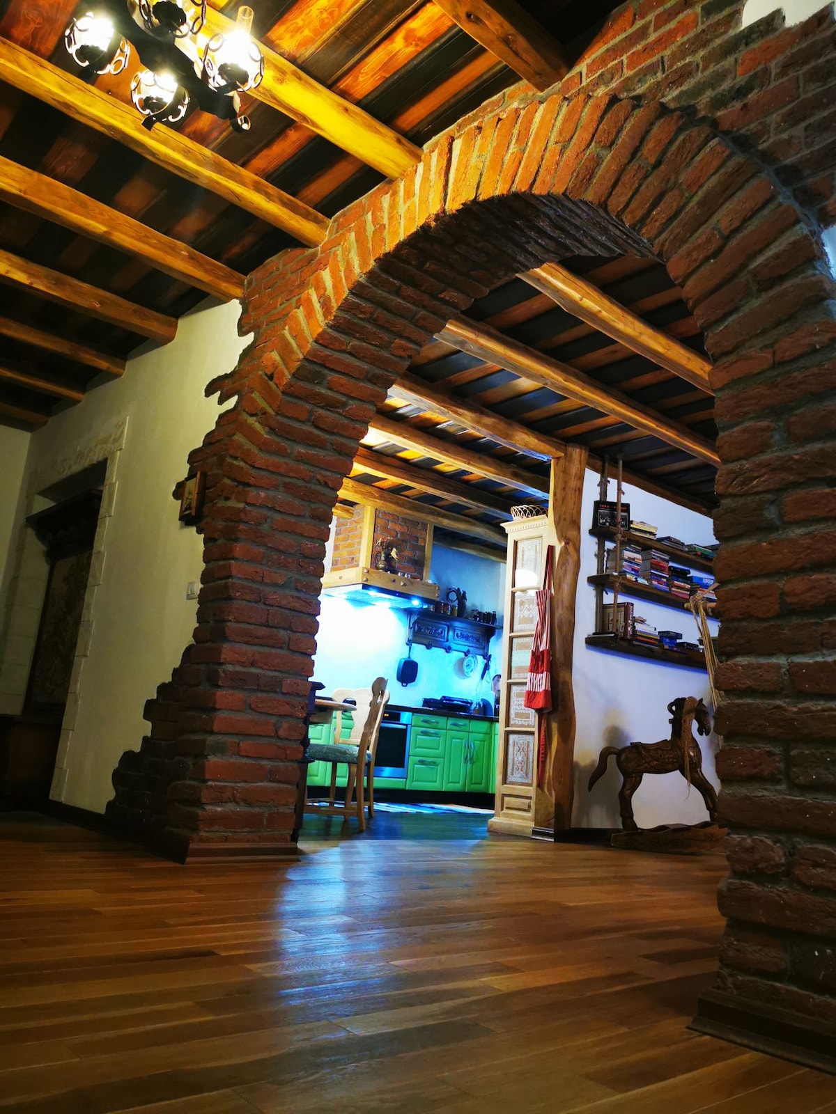 A brick archway leads into a well-lit kitchen area, showcasing colorful cabinetry and wooden furnishings. The space features exposed wooden beams along the ceiling and a warm wooden floor that enhances the cozy ambiance.
