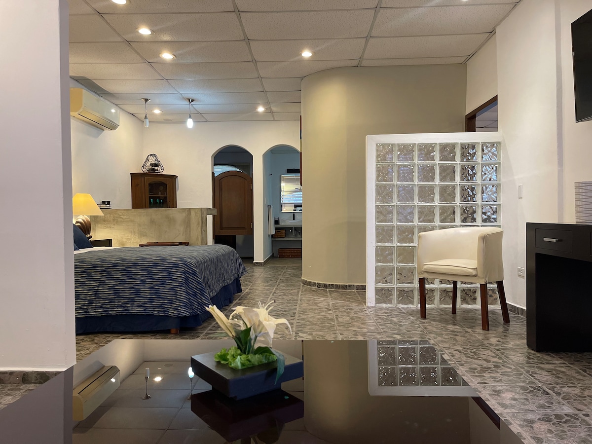 A spacious studio apartment features a comfortable bed dressed in blue linens, with a living area that includes a small round table and chair. Natural light enters through a glass block partition. Adjacent kitchen amenities and modern decor are visible, creating a functional space.