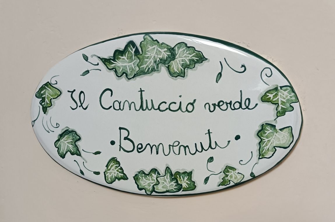 A decorative ceramic sign is displayed, featuring the text 'Il Cantuccio Verde' and 'Benvenuti' surrounded by hand-painted green ivy leaves. The sign's oval shape offers a warm welcome to guests upon arrival.