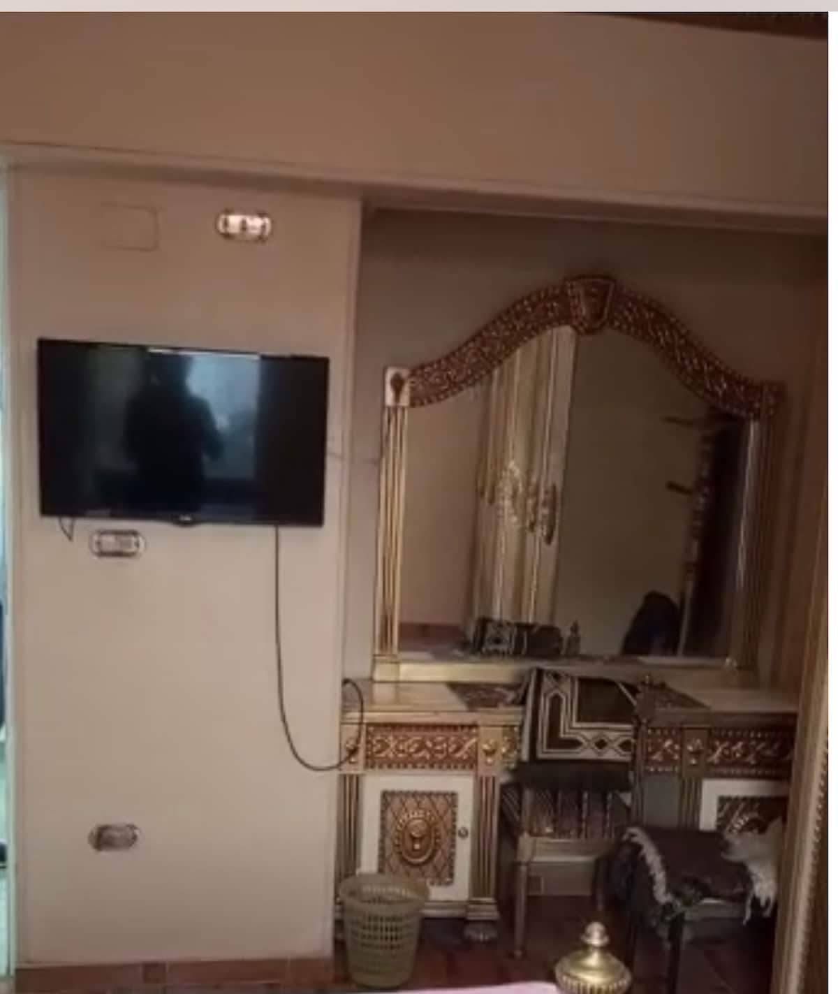 A wall-mounted television is positioned in front of an ornate vanity with an intricately designed mirror. The vanity is accompanied by a simple chair, and a small basket is placed nearby. Light is softly filling the room, creating an inviting ambiance.