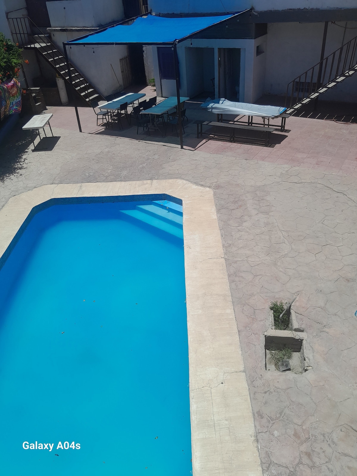 A clear blue swimming pool is framed by a light-colored deck. In the background, a shaded seating area with multiple tables features a blue canopy. A staircase is visible alongside the pool, leading to the upper level of the property.