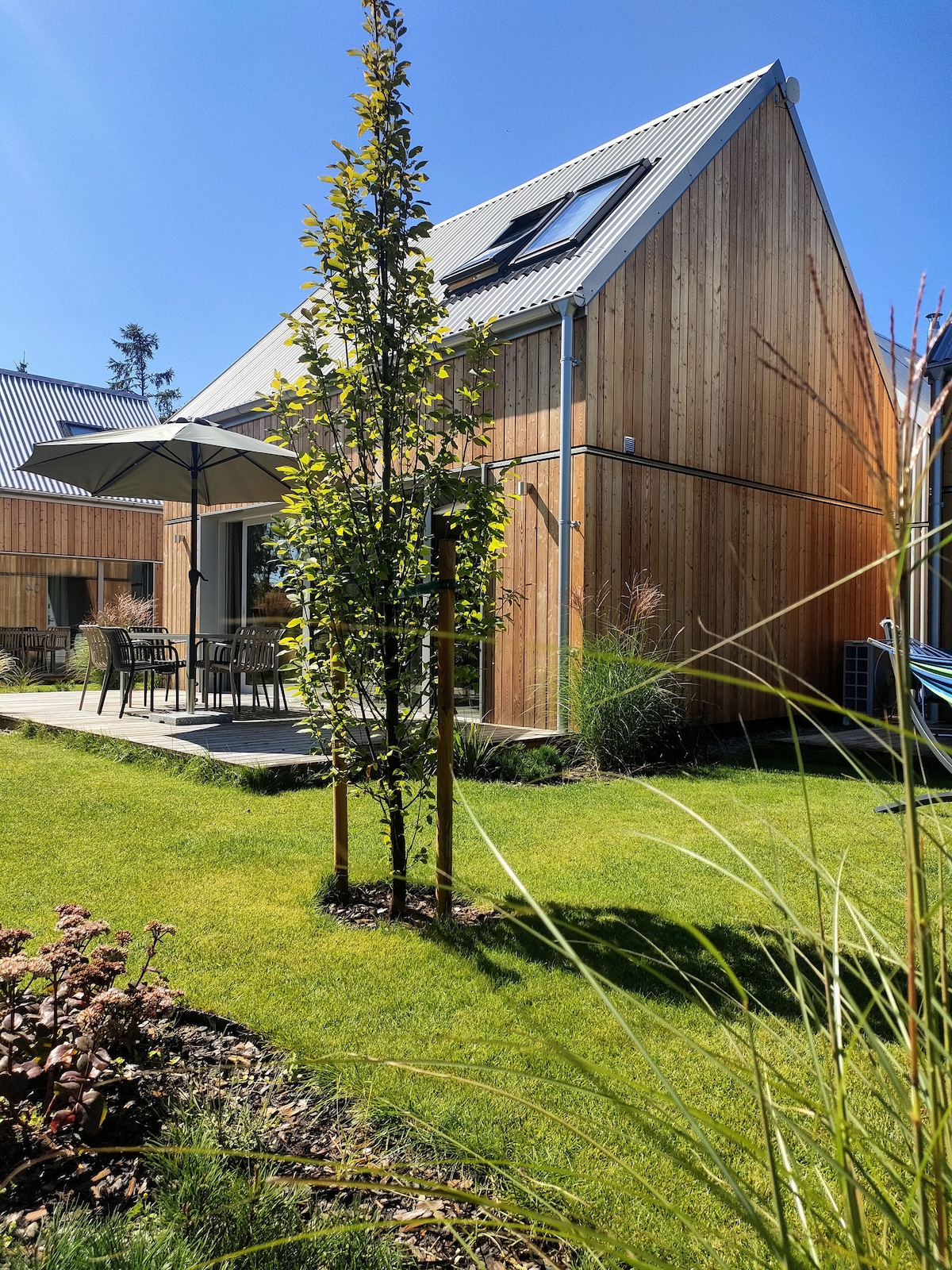 A modern wooden structure is depicted, featuring natural materials and minimalist design. A shaded patio area with outdoor furniture is visible. Lush green grass surrounds the home, complemented by carefully maintained shrubs and a young tree, enhancing the tranquil outdoor setting.