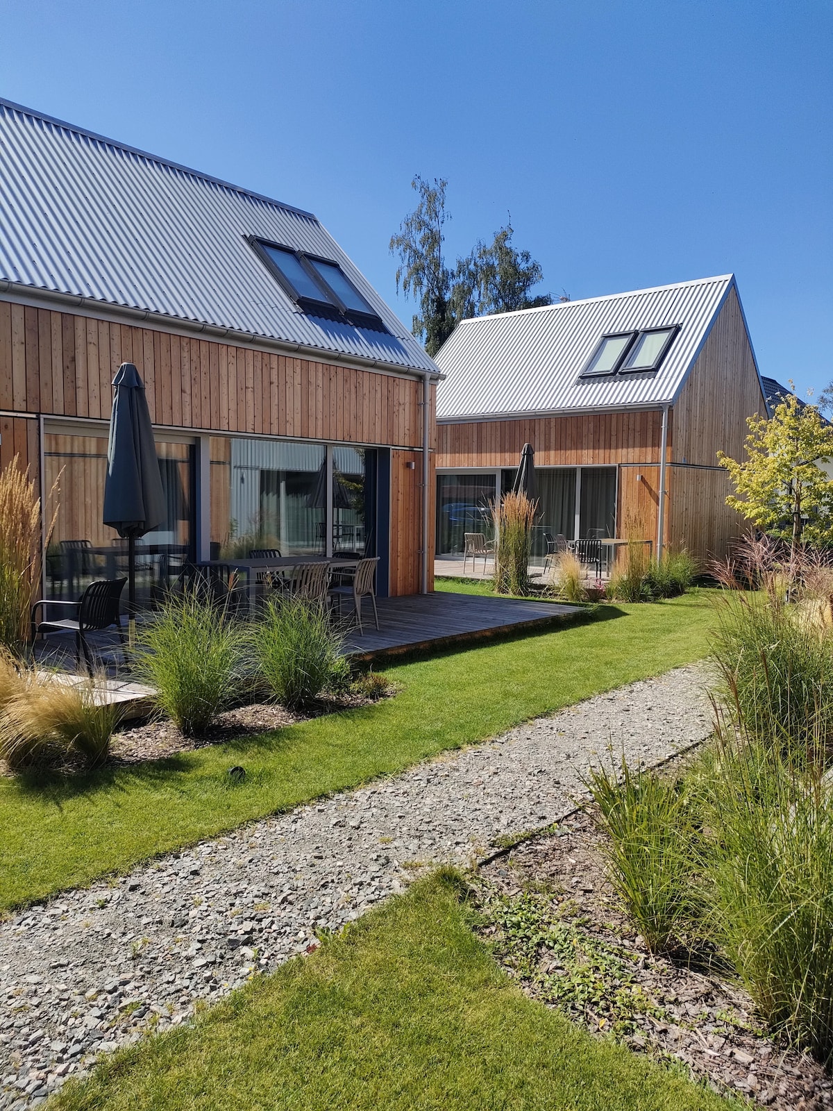 Two minimalist houses are positioned side by side, featuring wooden facades and large glass windows. A well-maintained garden with green grass and decorative shrubs surrounds the houses. Outdoor seating areas with black umbrellas can be seen on the spacious wooden terraces.