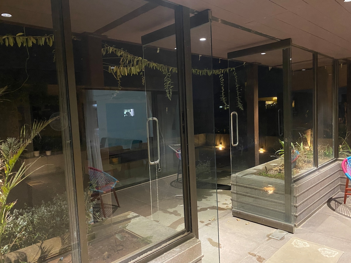 A glass entryway is framed by sleek metal doors, revealing lush greenery and soft lighting in the background. The outdoor area is enhanced by planters and subtle illumination, creating a serene evening ambiance.
