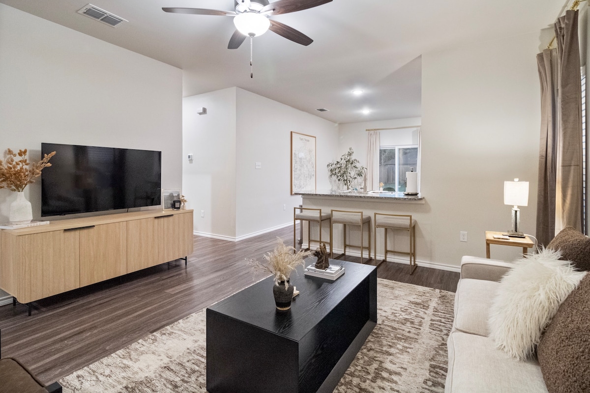 A spacious living area features a modern wooden TV unit with a large screen television. Light-toned walls complement the warm hardwood flooring. A cozy sofa is paired with a dark coffee table, and a dining area with bar seating is visible in the background.