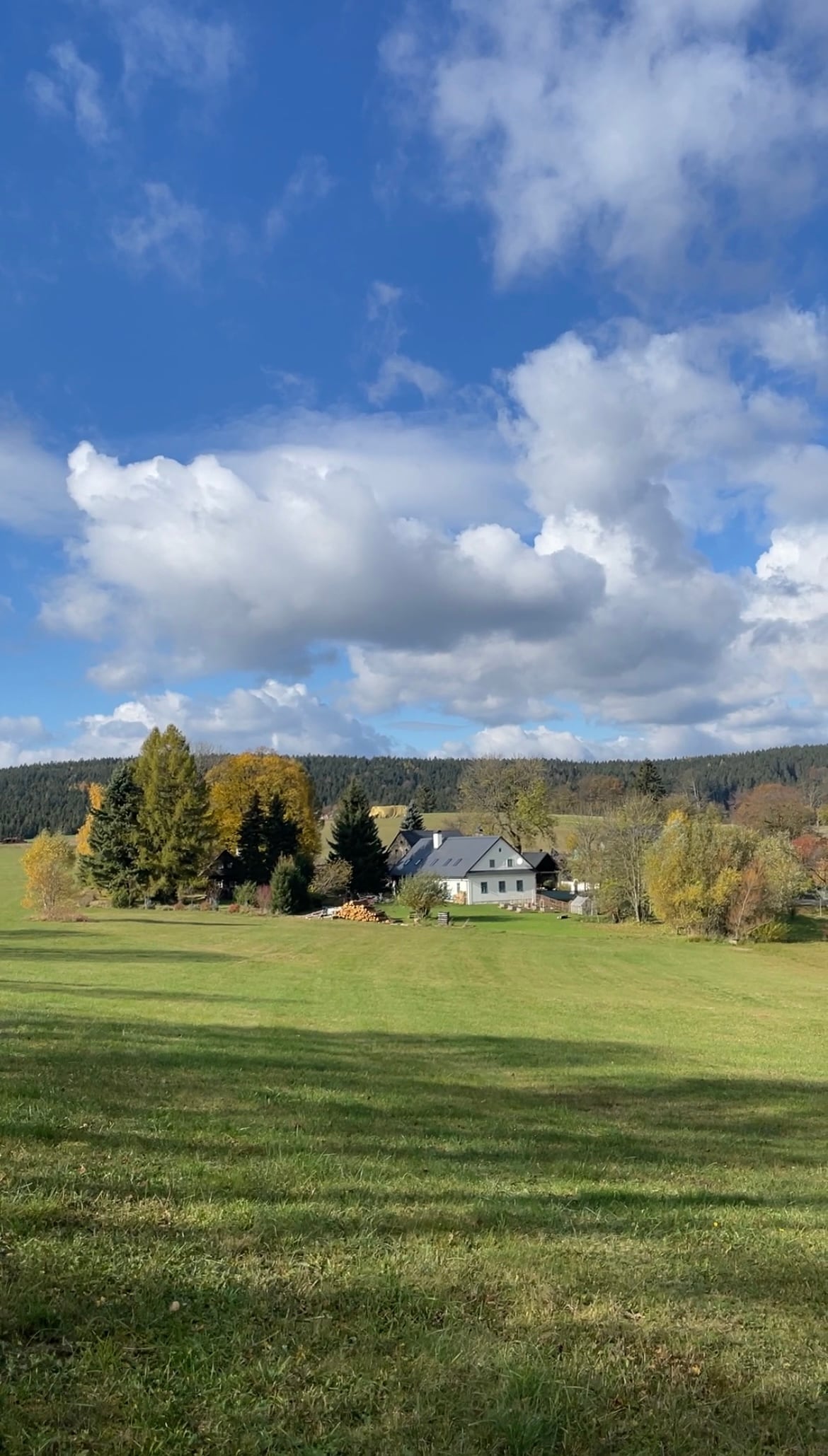 A spacious green field extends towards a charming traditional house, framed by colorful autumn trees. The backdrop features rolling hills under a partly cloudy sky, highlighting the peaceful rural setting. The vibrant greenery contrasts with the warm hues of the surrounding foliage.