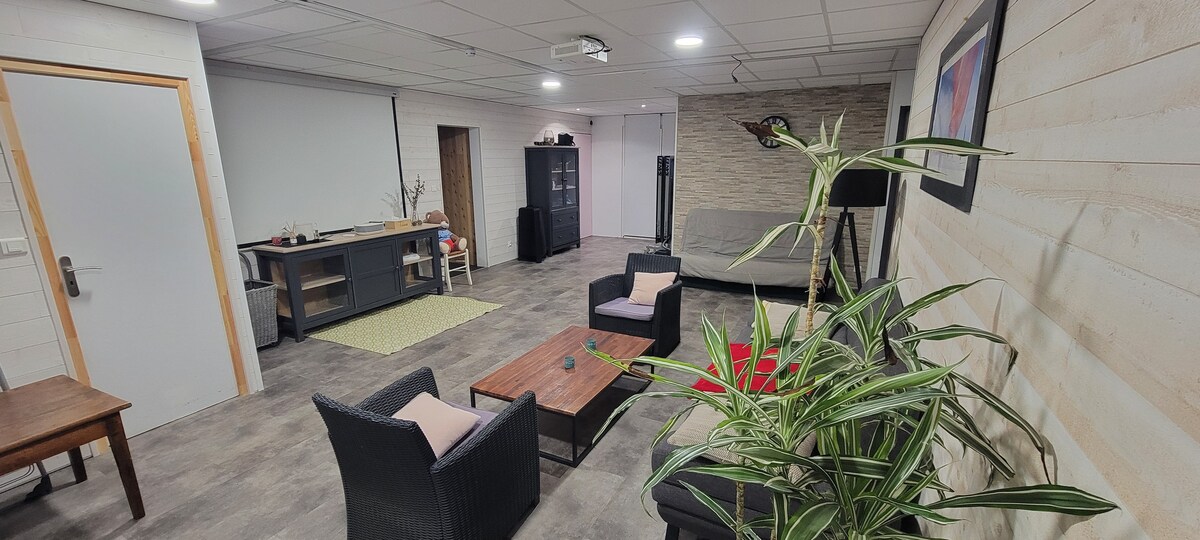 A spacious living area features seating arrangements that include a sofa and several chairs. A coffee table is positioned centrally, with potted plants adding a touch of greenery. A projection screen is visible at the front, enhancing the space's functionality.