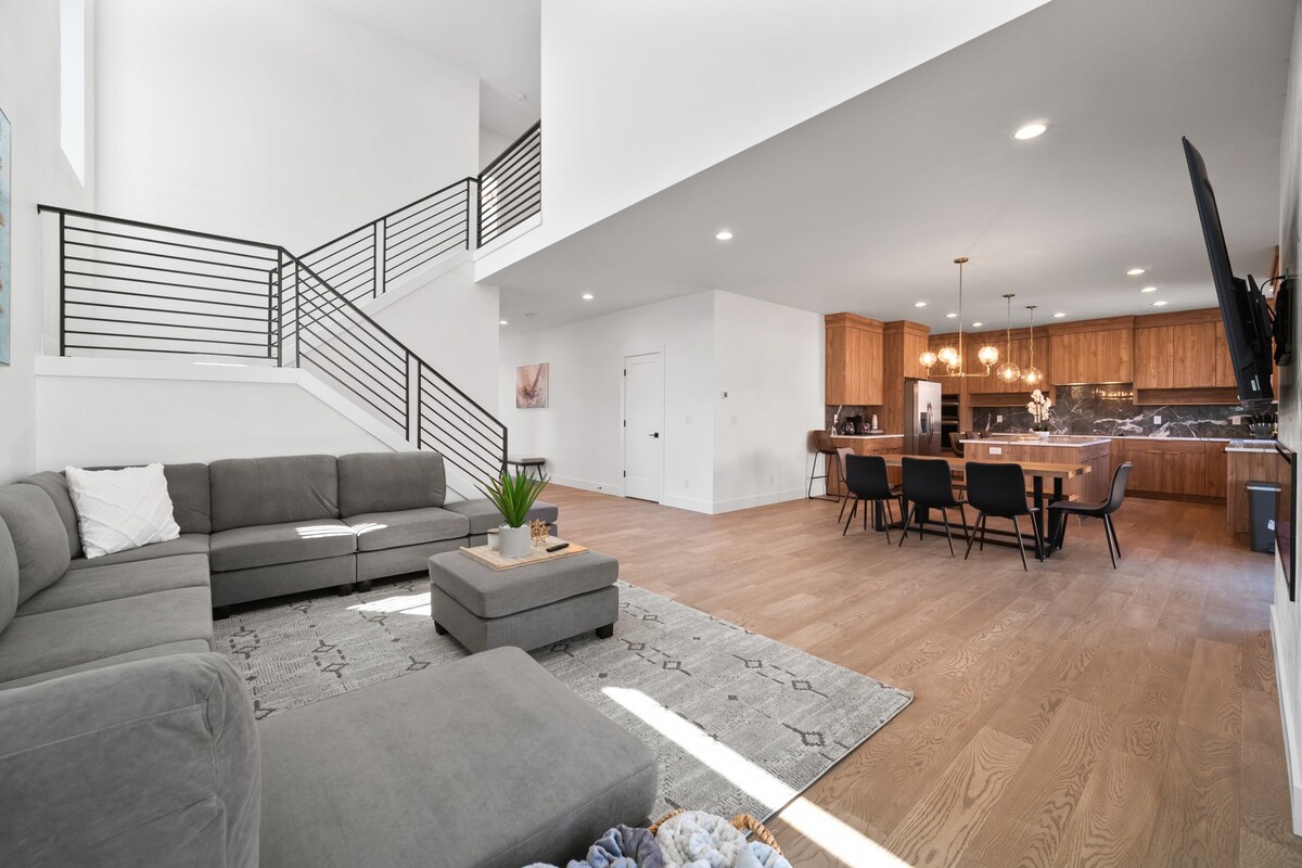 An open-concept living area showcases a spacious sectional sofa and a stylish coffee table atop a light-colored rug. Natural light floods the space through large windows, illuminating the dining area and modern kitchen in the background, featuring wood cabinetry and elegant pendant lighting.