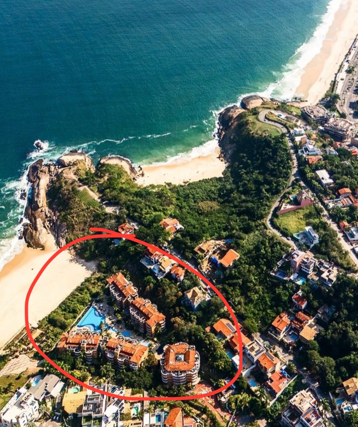 An aerial view captures the resort-style condominium complex nestled among lush greenery, with immediate access to the sandy beach. The coastline stretches alongside the turquoise waters, and the surrounding area is characterized by a blend of residential and natural landscapes.