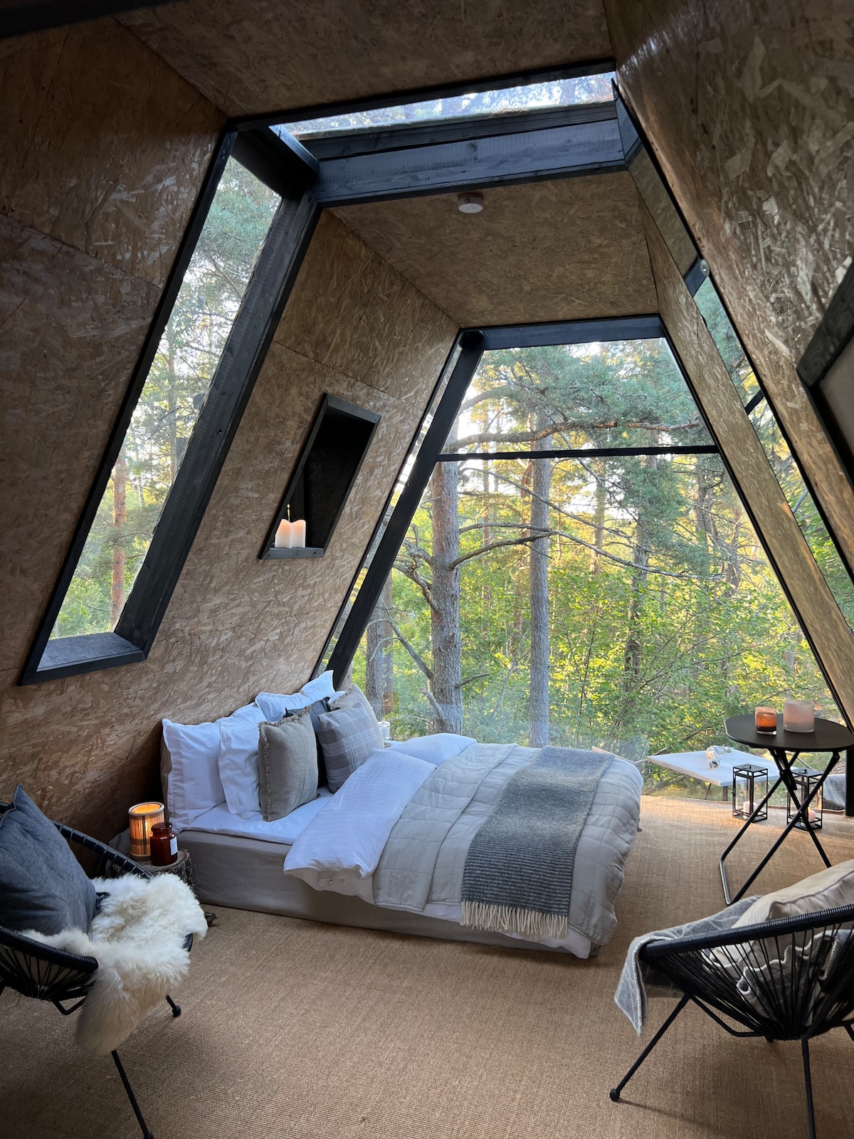 A cozy sleeping area is featured within the A-frame, showcasing large windows that allow natural light to fill the space. A spacious bed is neatly arranged with soft linens, complemented by a warm throw. Two inviting chairs and a small table are positioned nearby.