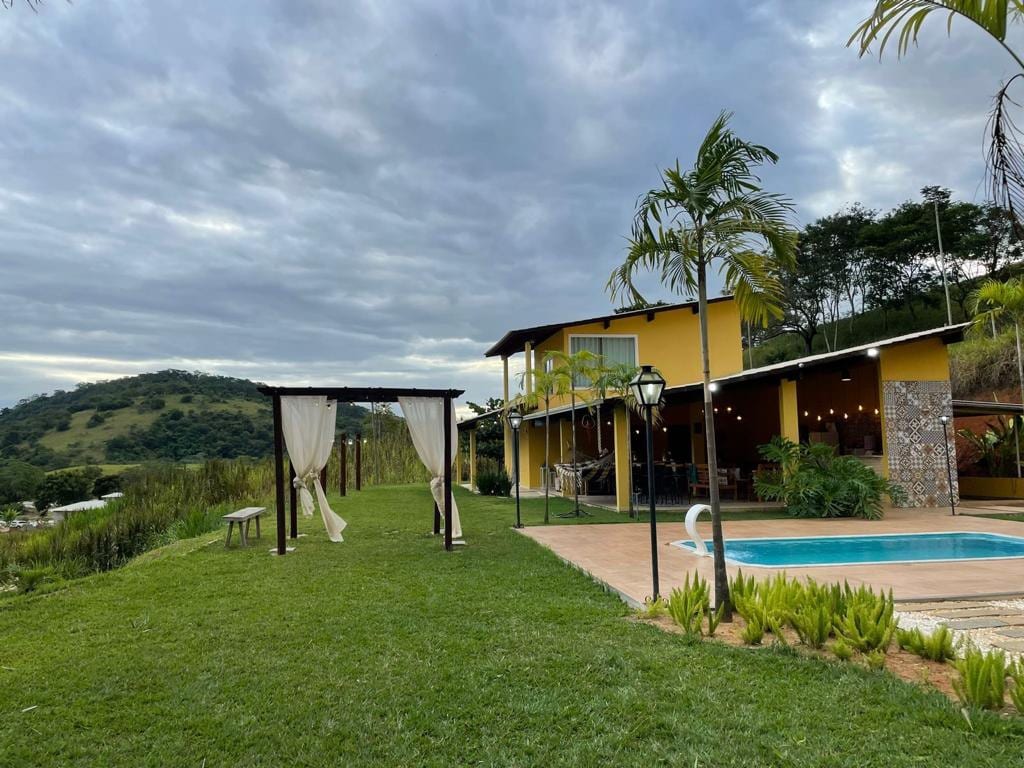 An inviting outdoor area features a well-maintained lawn, a swimming pool, and elegant draped canopies. The two-story yellow house is positioned in the background, surrounded by greenery and hills, providing a serene setting for relaxation and leisure.