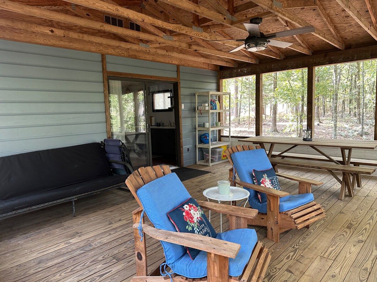 A spacious screened-in porch features wooden flooring and a ceiling fan. Two blue chairs with decorative cushions are positioned around a small table. A picnic table is seen in the background, and a black sofa can be spotted near the entrance to the cabin.