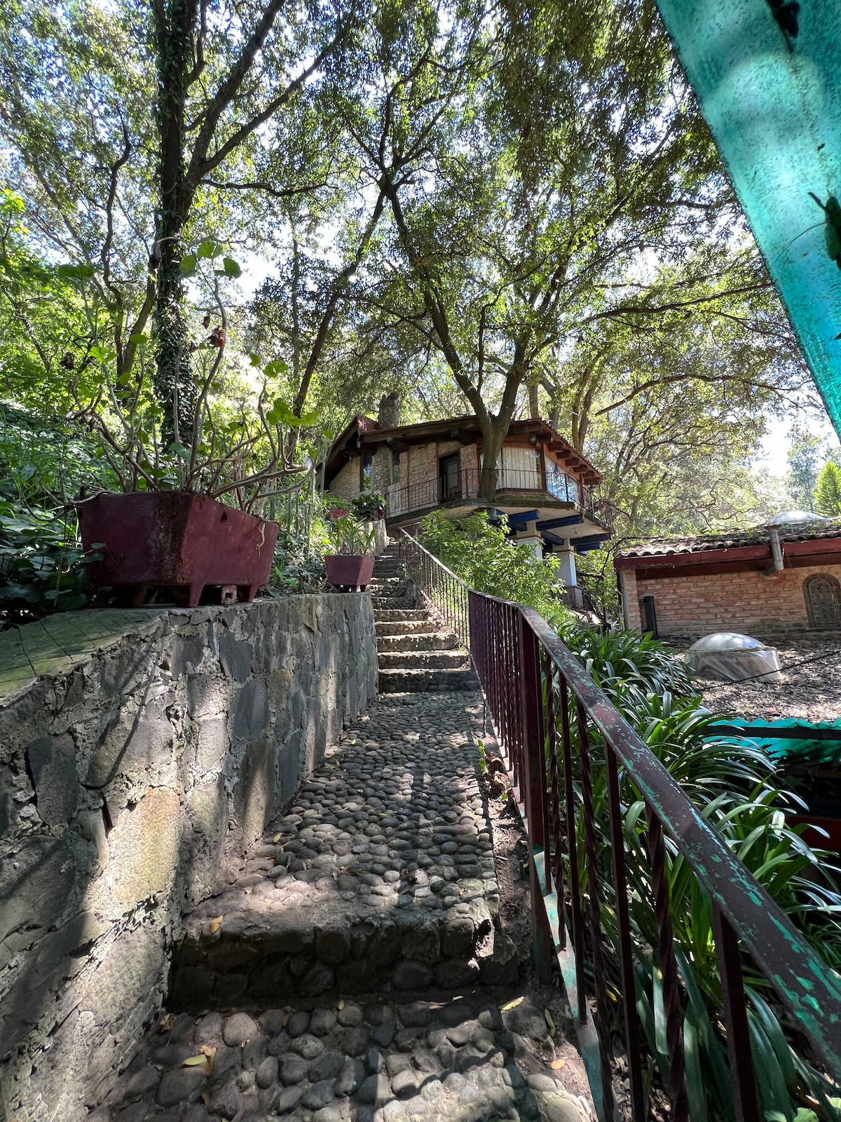 A stone pathway leads upward, flanked by lush greenery and large planters. A charming cabin is visible at the top, surrounded by trees that provide shade. The steps are bordered by a wrought-iron railing, enhancing the natural setting of the property.