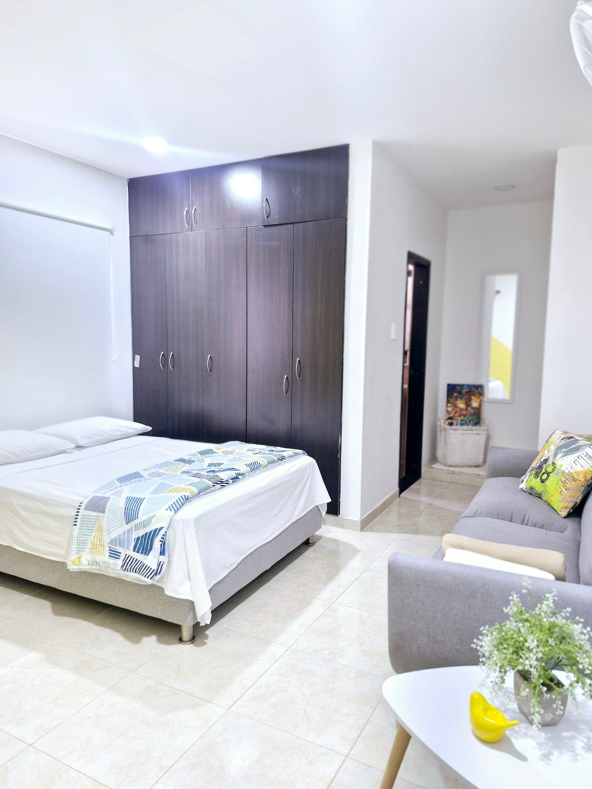 A well-lit room features a spacious bed with a light-colored blanket and a built-in closet with dark wood doors. A comfortable gray sofa is positioned next to the bed, and a small decorative mirror is visible on the wall opposite the entrance.