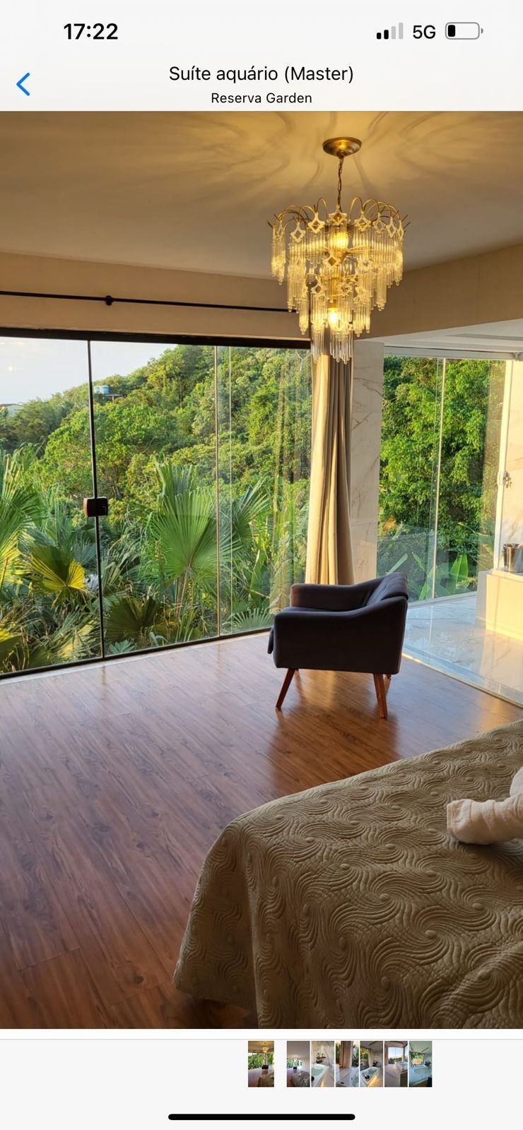 A well-lit suite features large glass doors providing views of lush greenery. A stylish chair sits beside the bed, with elegant decor including a chandelier above. The warm wooden flooring complements the neutral-toned bedding, creating a serene space for relaxation.