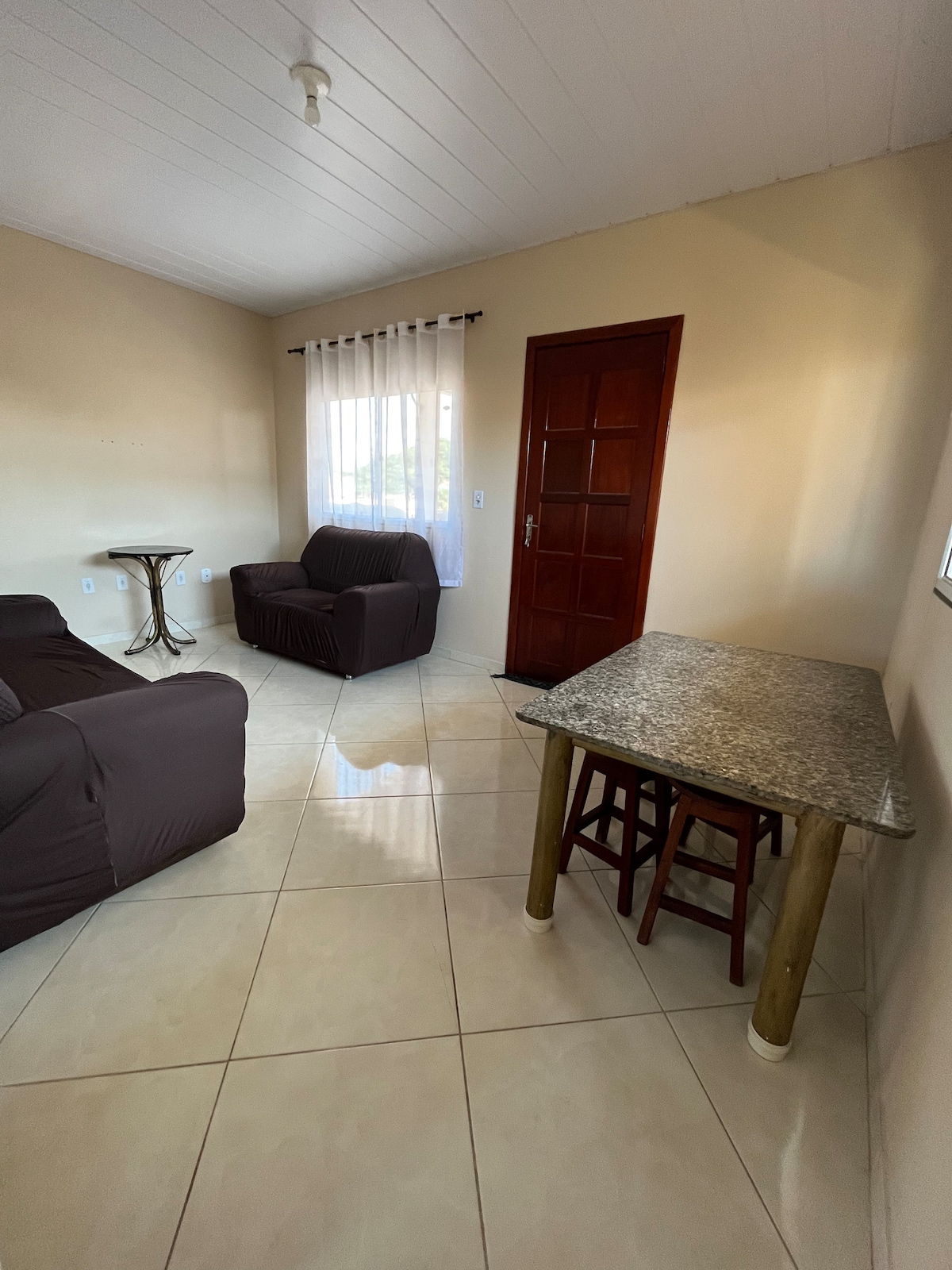 The living area features a pair of comfortable armchairs and a small dining table with wooden legs. Natural light enters through a window dressed with sheer curtains, illuminating the tiled floor and warm beige walls.