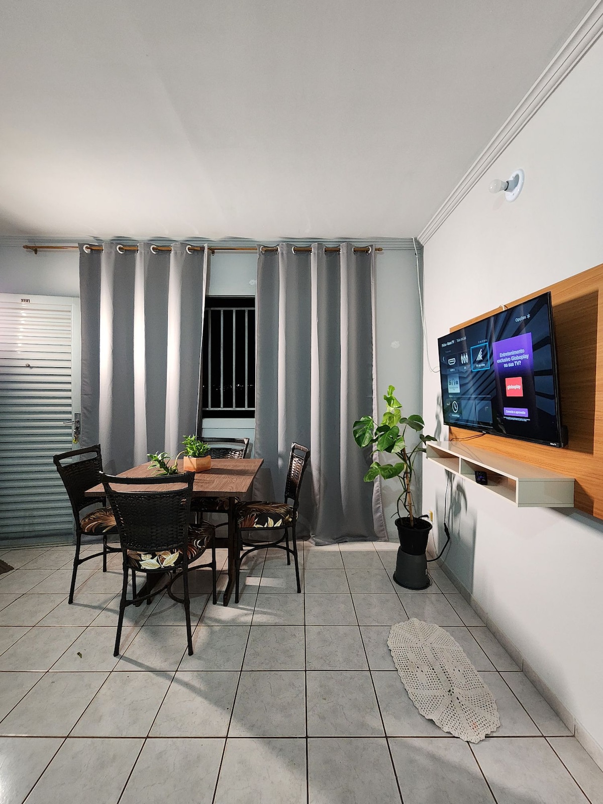 A dining area is shown with a wooden table surrounded by four wicker chairs. A TV is mounted on the wall, and a potted plant adds a touch of greenery. Light-colored tiles cover the floor, while curtains provide a soft backdrop.