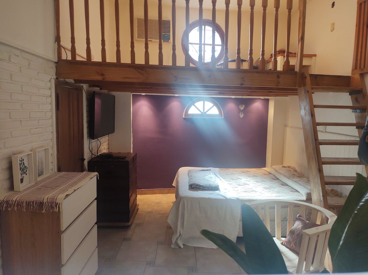 A spacious room features a comfortable bed positioned beneath a lofted area. Natural light flows through a round window, highlighting the purple accent wall. A dresser and TV are present, accompanied by a staircase leading to the upper level, creating an open and airy atmosphere.