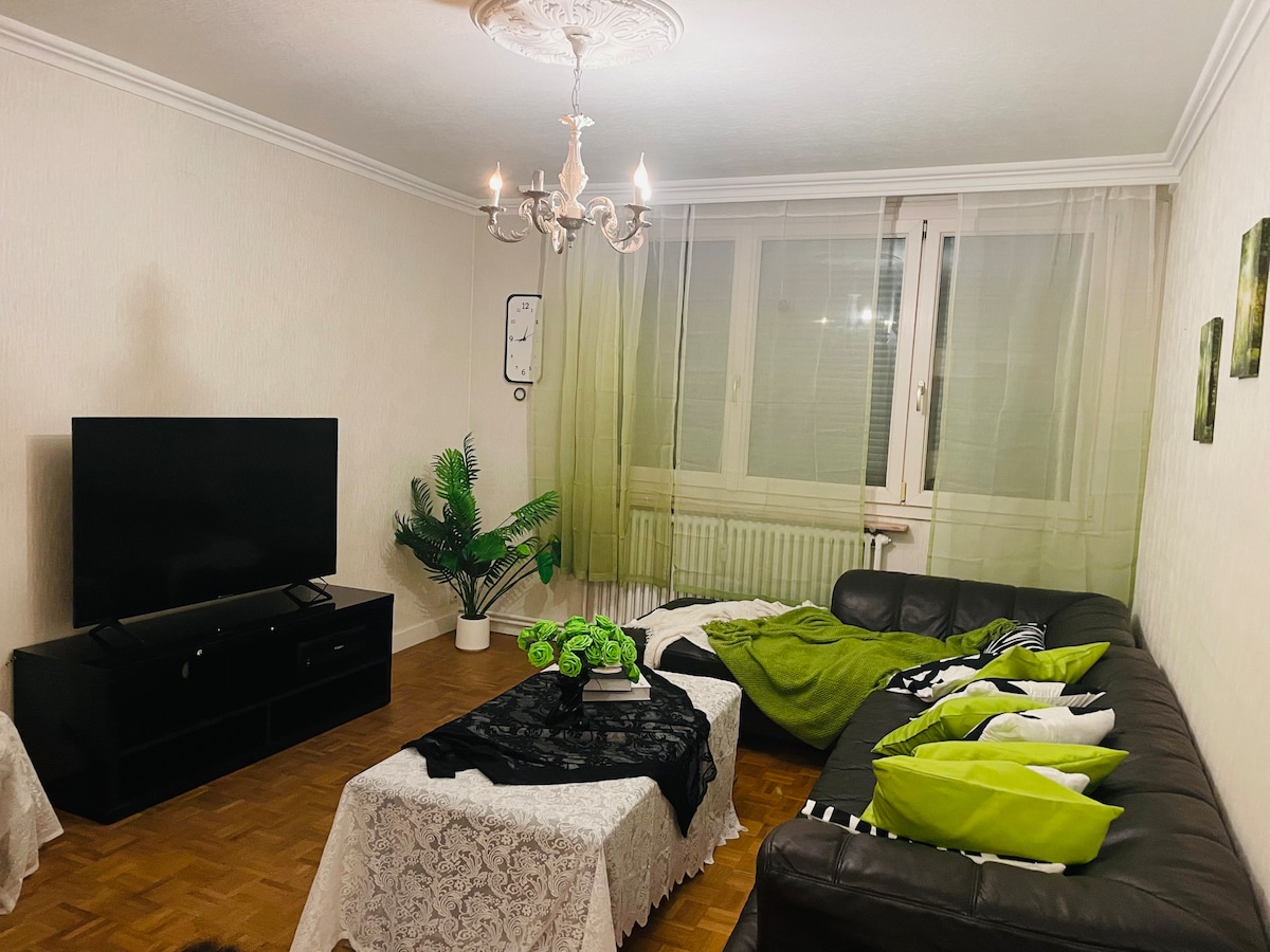 A welcoming living area is presented with a dark leather sofa adorned with green and white cushions. A coffee table covered in a lace tablecloth sits in front of a mounted television. Sheer green drapes allow soft light to filter in from the large windows.