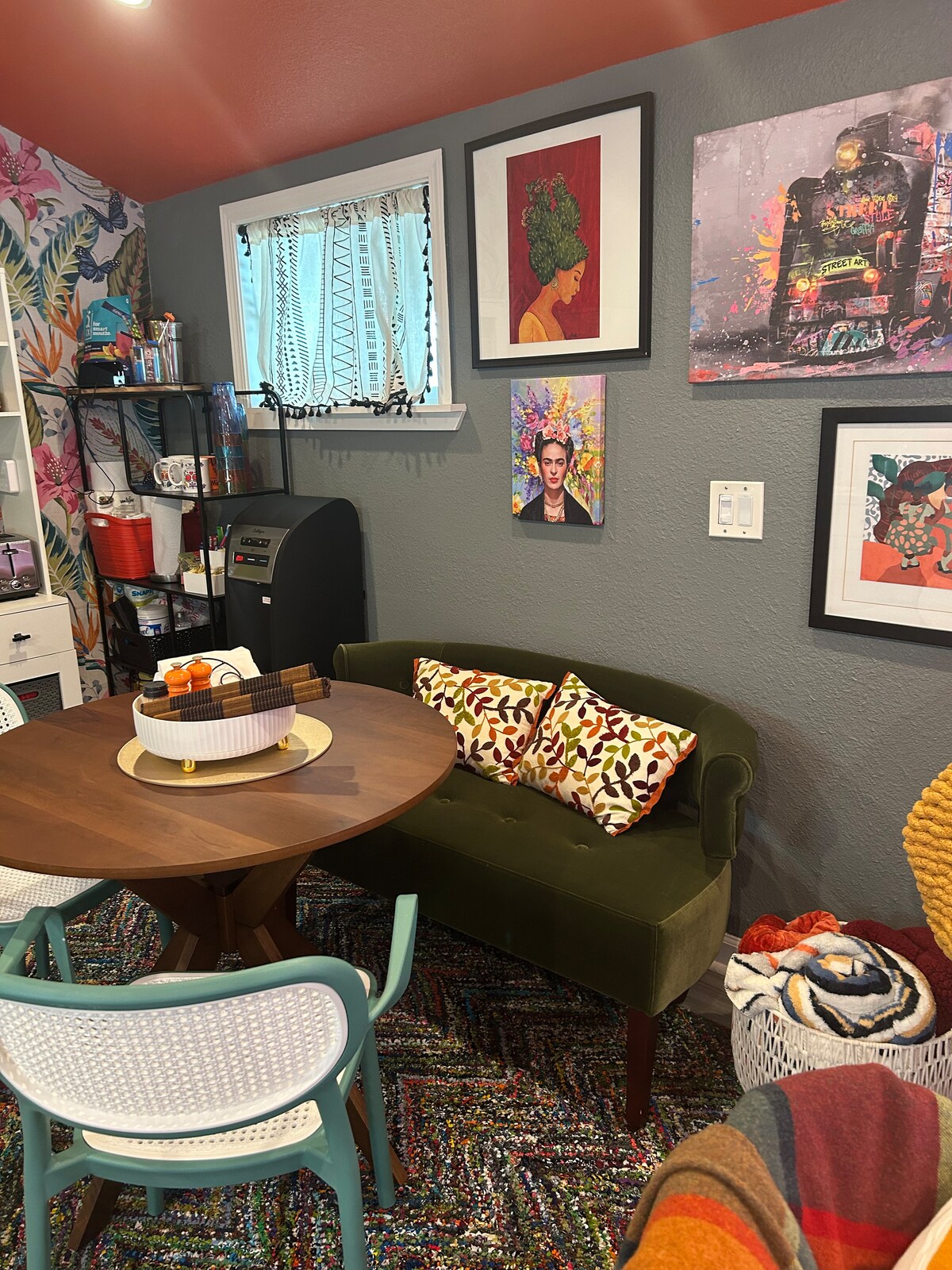A vibrant seating area features a round table surrounded by various chairs, including green and white options. Colorful artwork adorns the walls, and patterned cushions are placed on the green sofa. A cozy rug adds texture to the inviting space.
