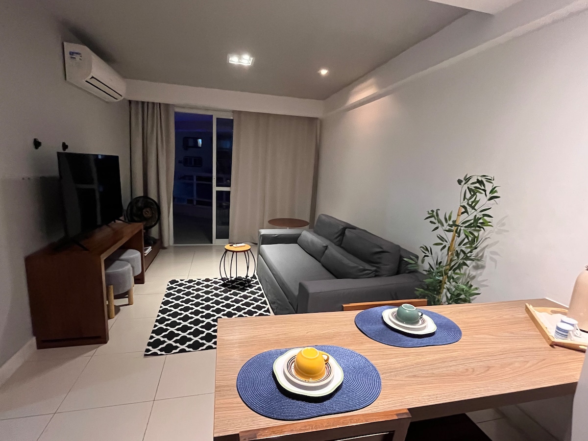 A modern living space features a comfortable gray sofa positioned across from a mounted television. A small round table complements the seating area, while a dining table is set with dishware. Soft lighting enhances the ambiance, with curtains framing a balcony door.