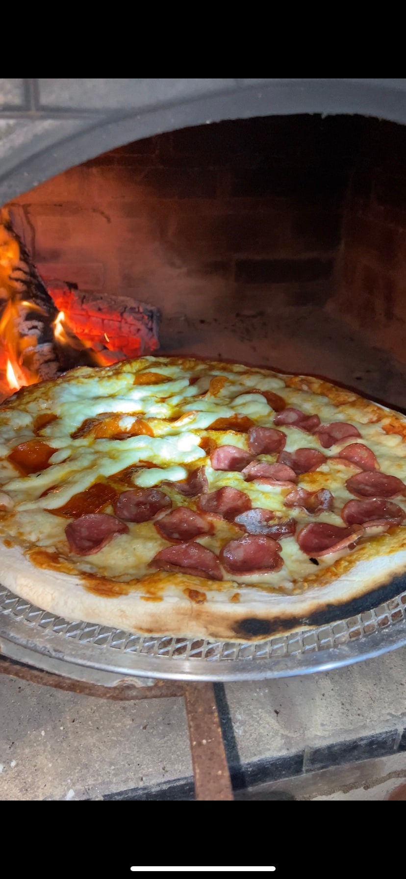 A pizza with a golden crust and toppings of cheese and pieces of sausage sits on a rack inside a wood-fired oven. The glow of the fire in the oven creates a warm ambiance, with flames visible in the background.