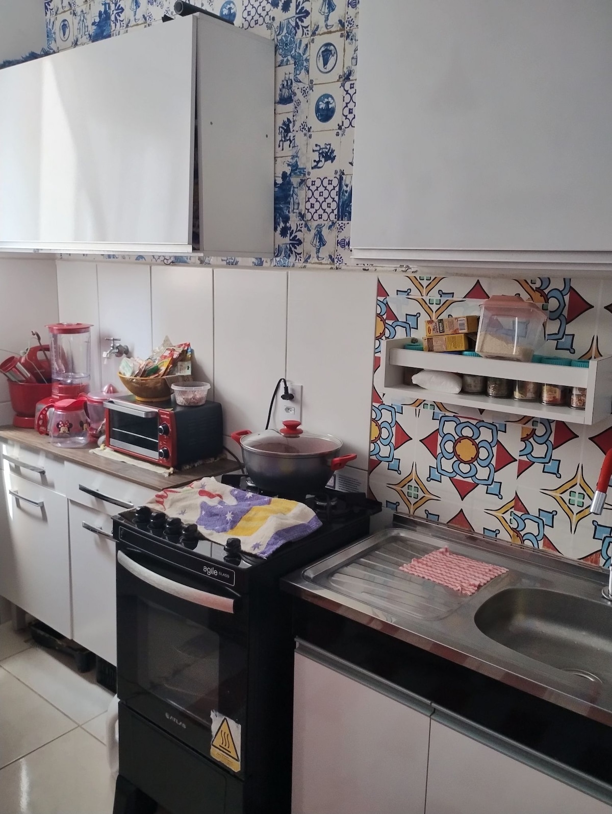 A compact kitchen features white cabinetry complemented by a patterned backsplash. A stove is positioned beside a stainless steel sink, while small appliances and kitchenware are organized on the counters. Natural light illuminates the space, highlighting its functional design.