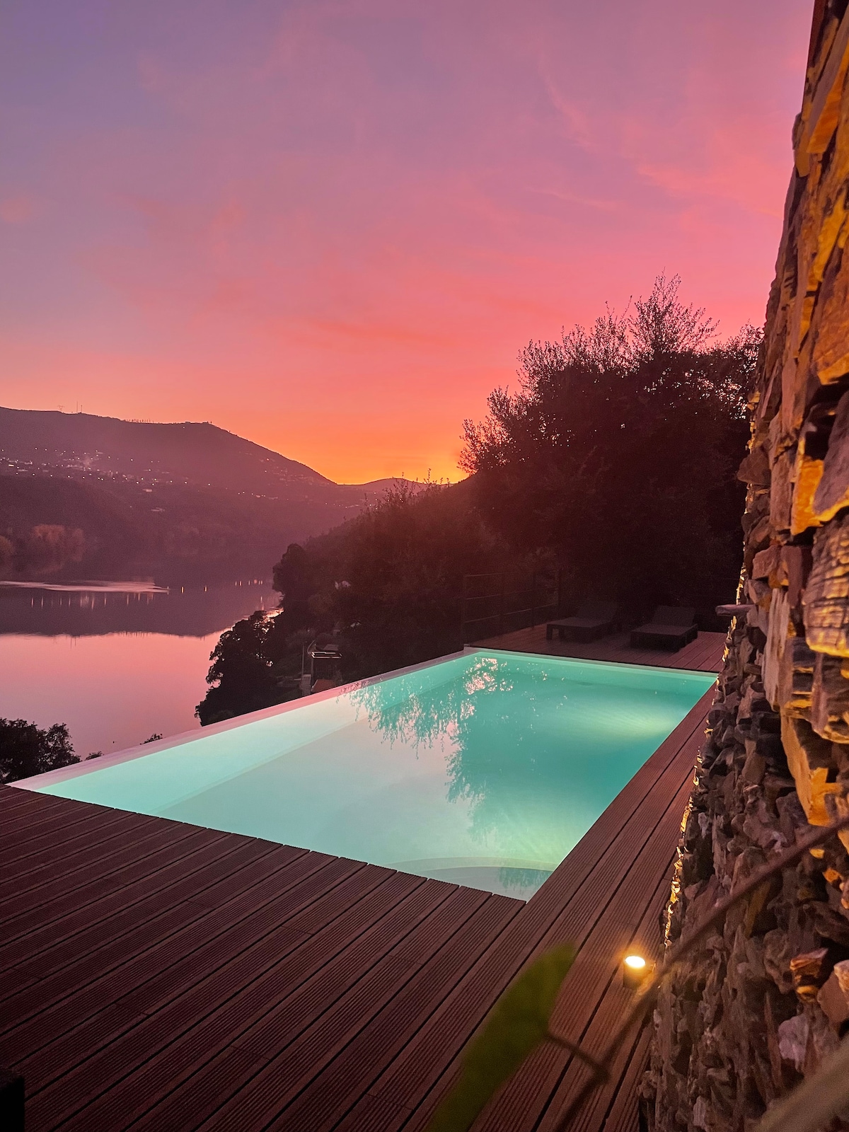 The image captures a clear saltwater pool surrounded by a wooden deck, reflecting the vibrant colors of a sunset. The landscape features rolling hills in the background, and the tranquil River Douro is visible, enhancing the serene and secluded ambiance.