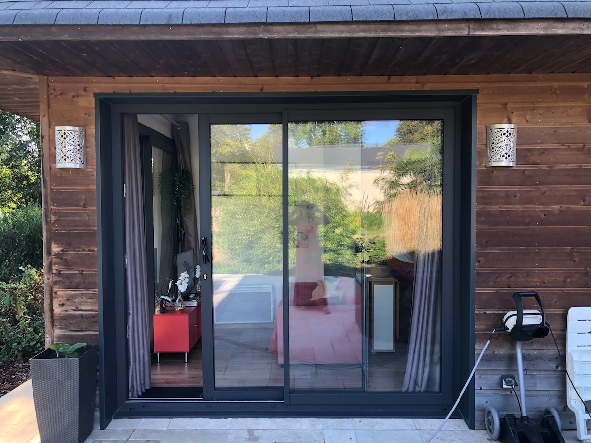 A contemporary entrance to a wooden structure showcases large sliding glass doors. The doors reflect the surrounding greenery, with hints of colorful furnishings visible inside. A small potted plant is positioned beside the door, adding to the inviting exterior.