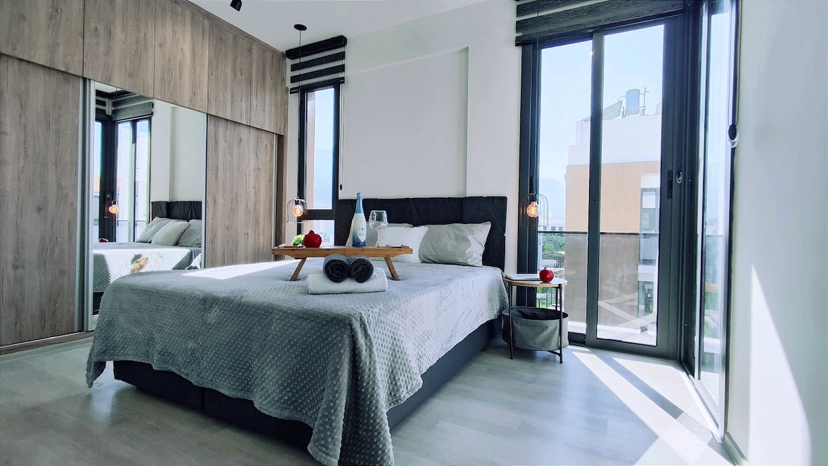A modern bedroom features a spacious bed topped with soft linens and neatly arranged towels. Large windows provide ample natural light and showcase views of the surrounding area. A stylish bedside table holds a refreshing drink, while a mirror reflects the contemporary design.