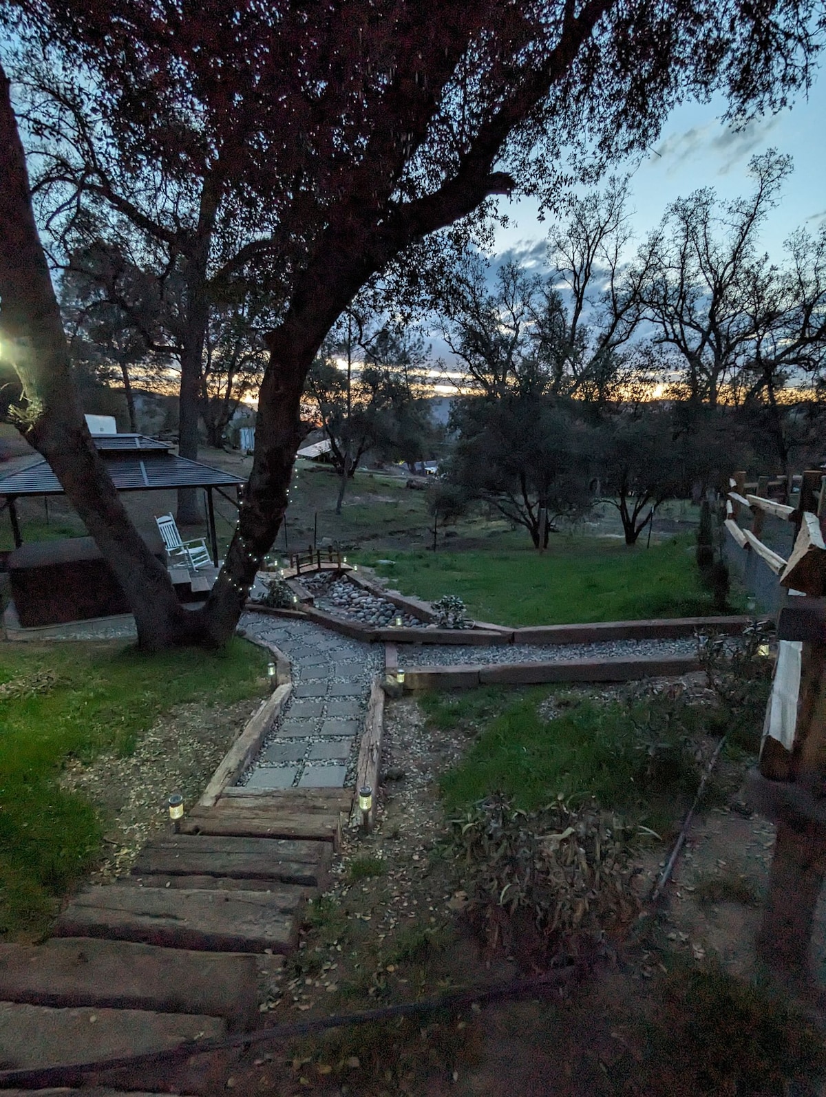 A winding pathway lined with smooth stones and bordered by grass leads through the property. Soft lighting from lanterns illuminates the path, creating a gentle glow amidst the surrounding trees. The horizon is visible in the background, hinting at a fading sunset.