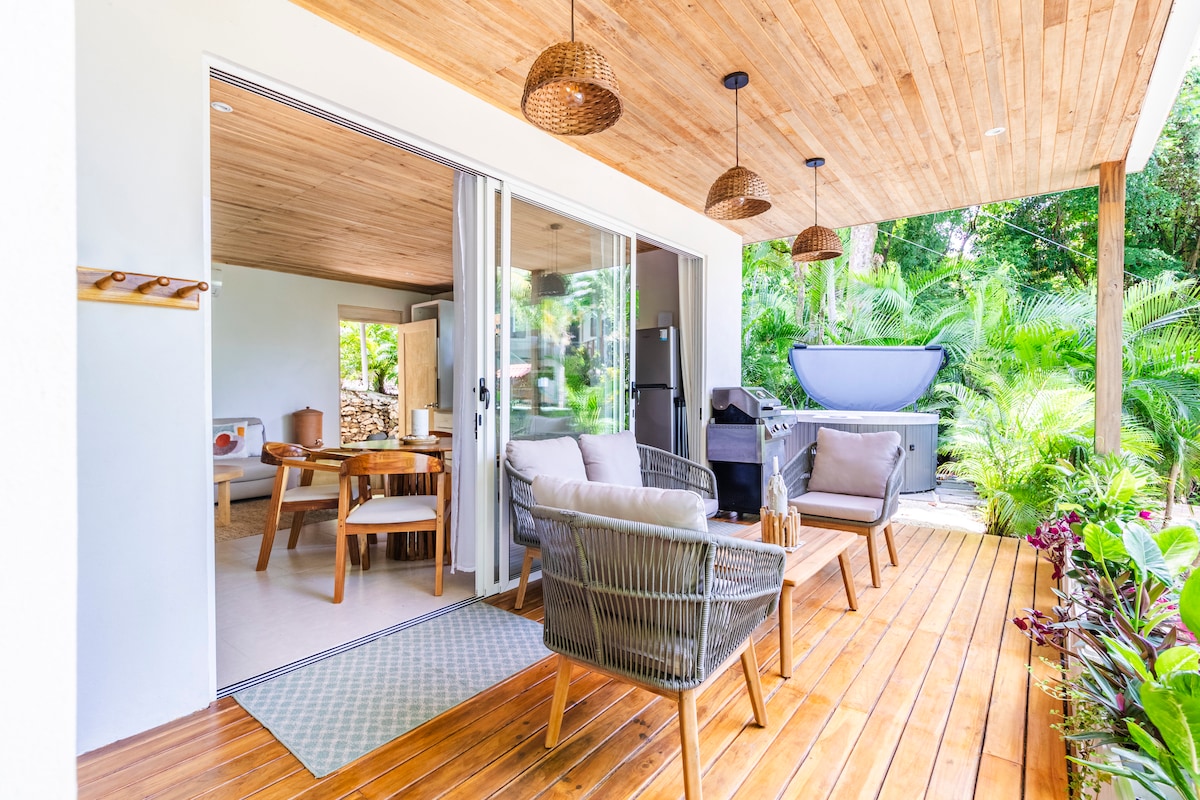 An outdoor terrace features natural wood flooring and a ceiling adorned with woven light fixtures. Comfortable seating arrangements with neutral cushions are positioned around a small table, inviting relaxation. Lush greenery surrounds the space, enhancing the connection to nature.