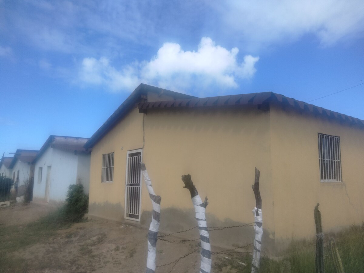 A single-story building with a sloped roof is featured, surrounded by open land. The exterior is painted a light neutral color, while windows are fitted with security bars. A small garden area is visible in the foreground, with a fence made of wooden posts and wrapped material.