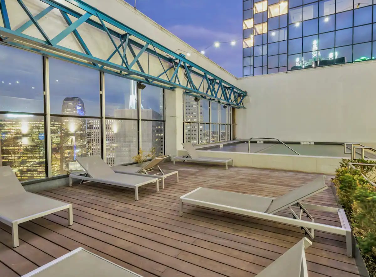 The rooftop area offers a spacious wooden deck with several lounge chairs positioned for relaxation. Large windows provide sweeping views of the city skyline, while string lights illuminate the space, creating a pleasant ambiance during the evening.