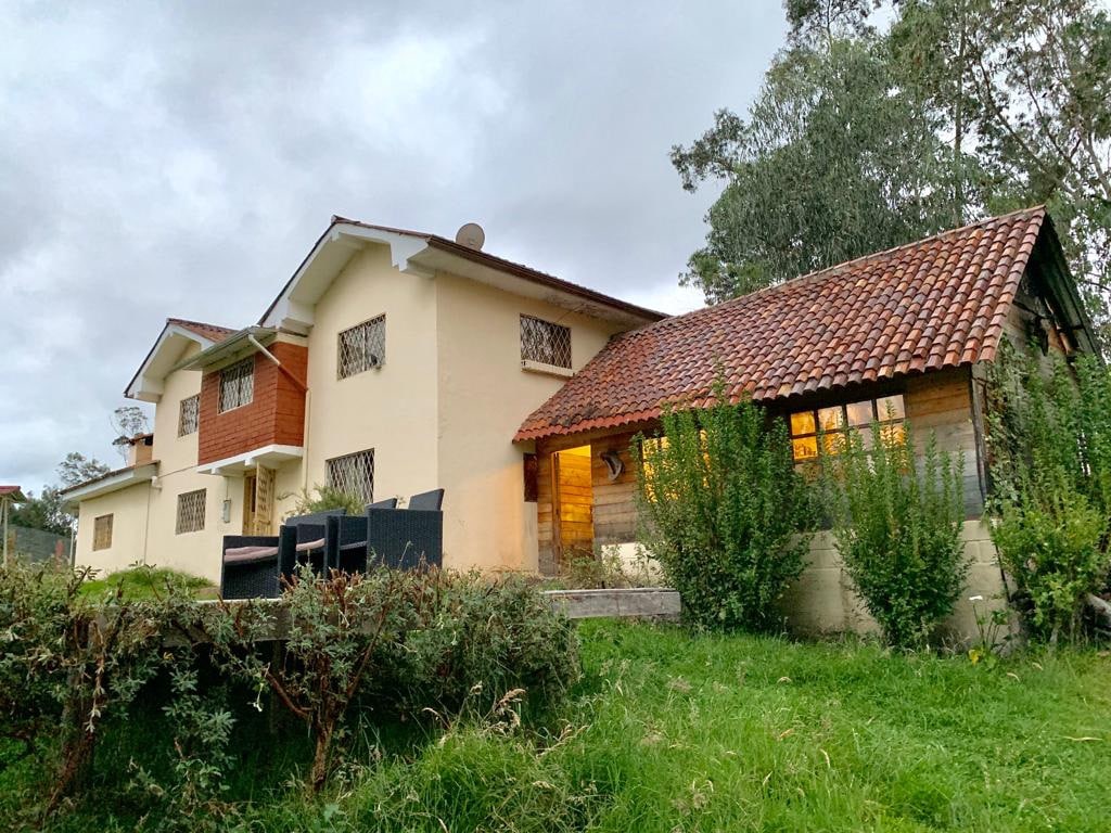 A two-story house is situated amidst greenery, featuring a red-tiled roof and multiple windows with grills. The exterior showcases seating arrangements on a patio, surrounded by well-maintained grass and shrubs, with trees in the background under a cloudy sky.