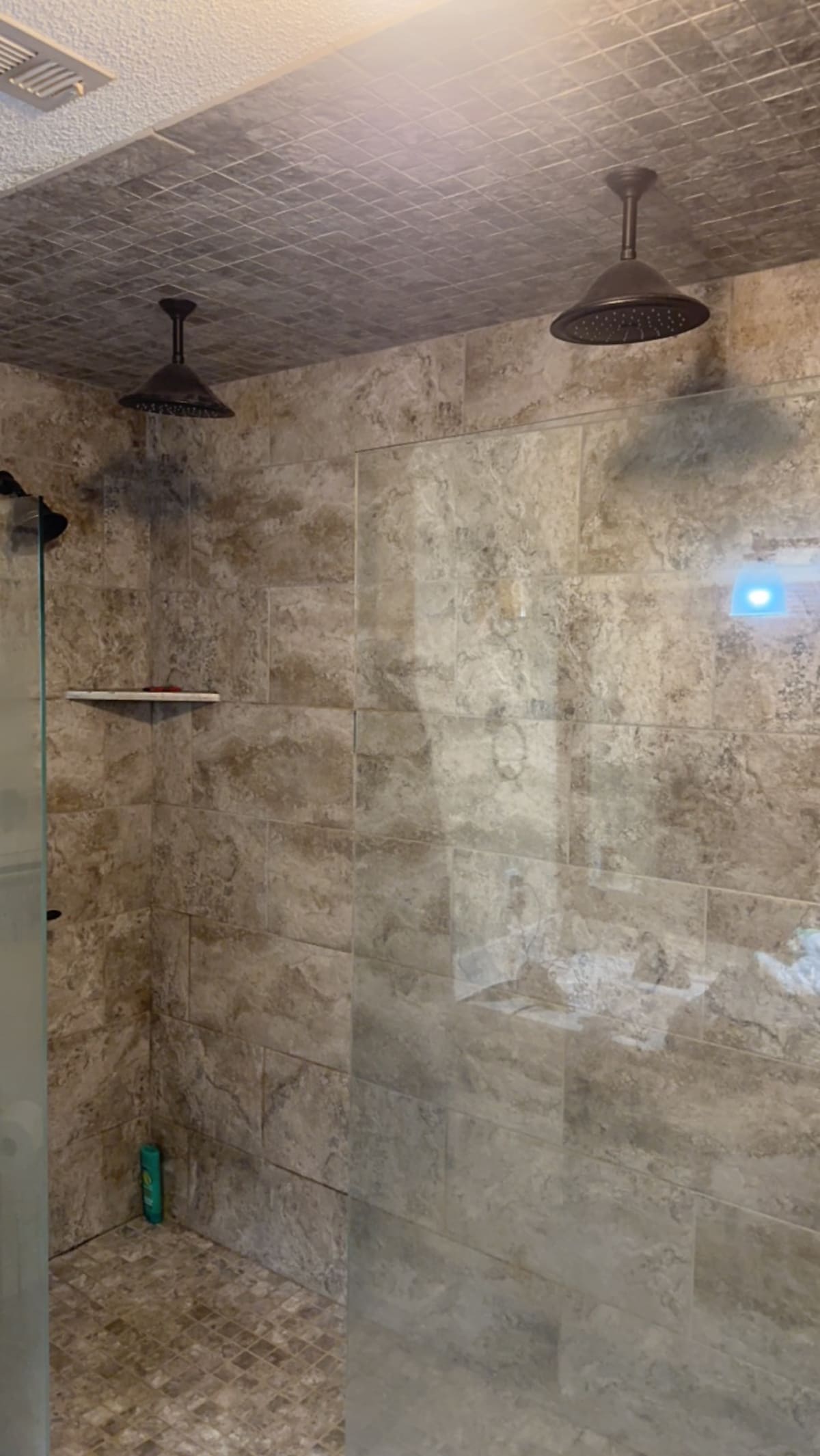 A spacious shower area features a modern design with stone-textured walls. Two shower heads are mounted from above, providing ample water flow. A glass partition offers an unobstructed view while a minimalist shelf is positioned for convenience. Light reflects off the tiled surfaces.