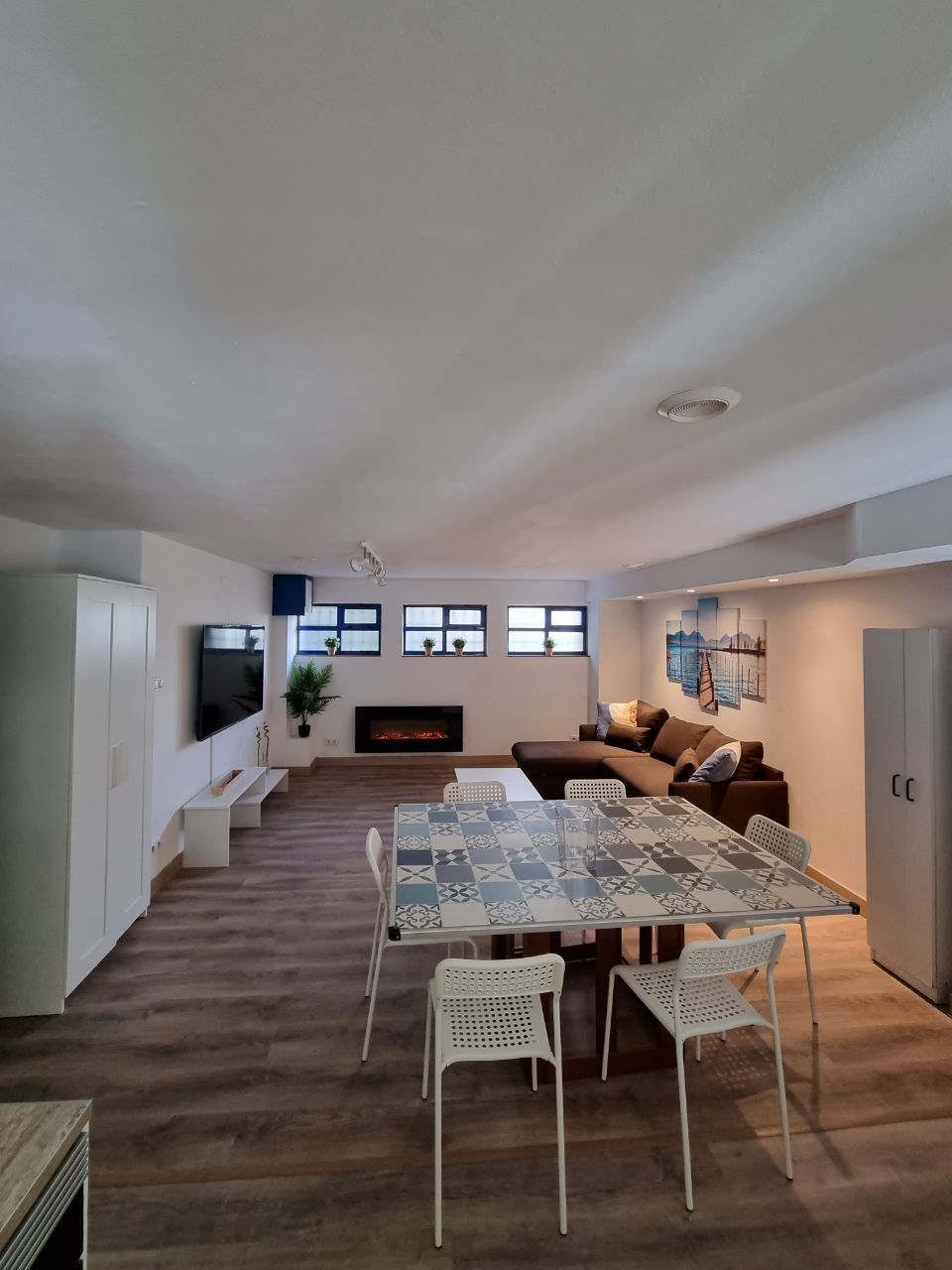 An open living area features a modern design with a cozy sofa facing a wall-mounted TV. A dining table with patterned tiles is surrounded by several white chairs. A sleek electric fireplace adds warmth, while large windows provide natural light.