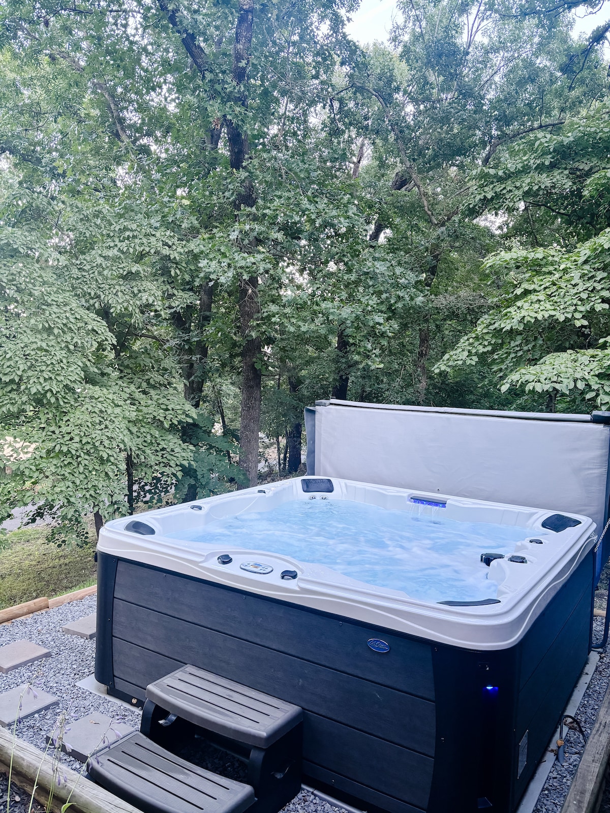 A six-person hot tub is positioned on a gravel patio, surrounded by lush greenery and tall trees. The tub features a white cover partially opened, revealing clear water ready for use. Steps lead up to the tub, providing easy access.