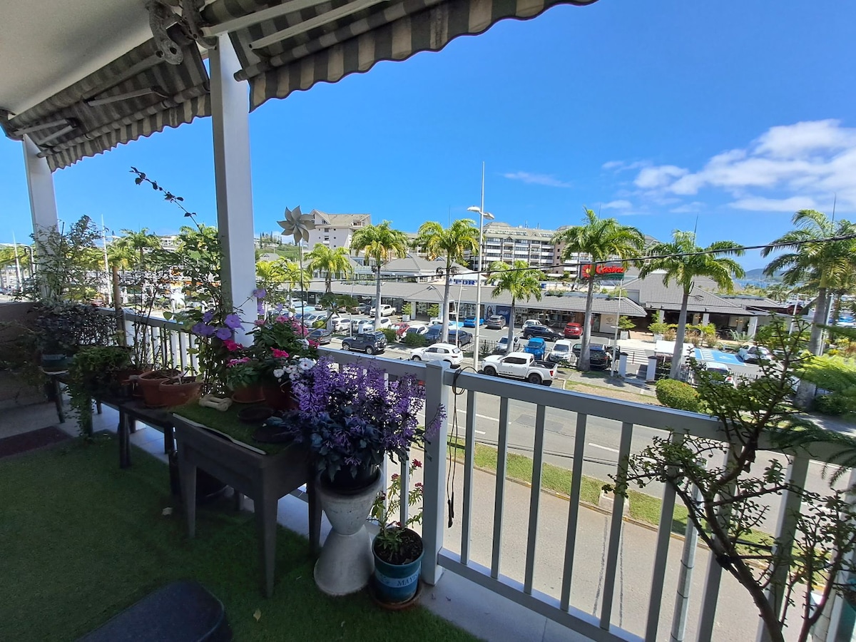 A balcony offers a view of palm trees lining a busy street. Potted plants, including colorful flowers, are arranged on a table, providing a touch of greenery. The scene captures a vibrant atmosphere with cars parked along the avenue and buildings visible in the background.