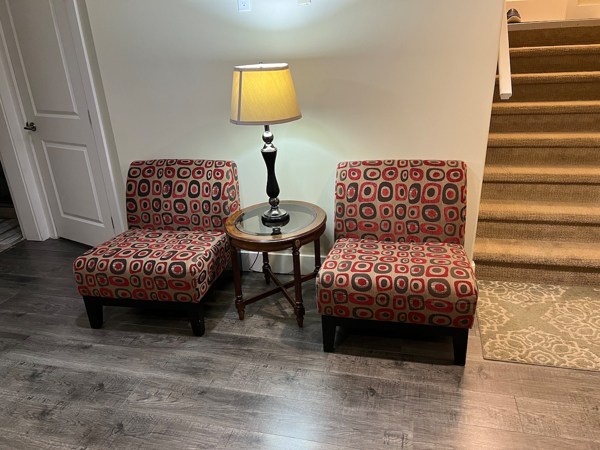 Two stylish accent chairs with a bold patterned fabric are positioned beside a small round table. A black lamp with a warm yellow shade stands on the table, offering soft lighting. A staircase with neutral colored carpeting is visible in the background.
