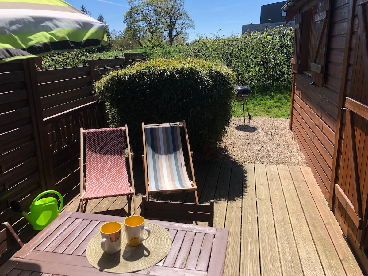 A wooden terrace features two lounge chairs positioned for relaxation under an umbrella. A small table with two mugs sits at the forefront. A BBQ grill is visible in the background, surrounded by a lush green hedge and trees, creating a serene outdoor space.