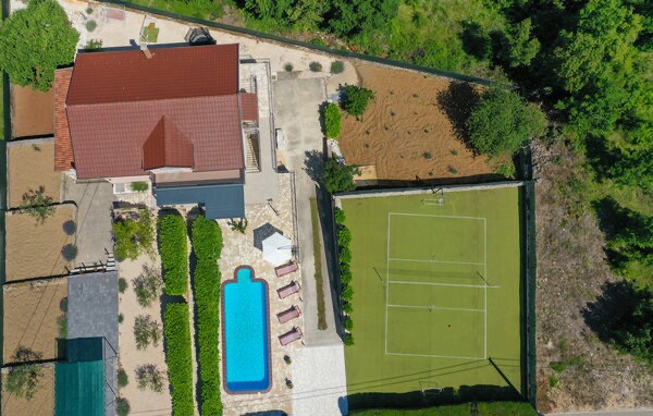 An aerial view of the property highlights a spacious outdoor area featuring a swimming pool surrounded by lounge chairs. A well-maintained tennis court is visible alongside a landscaped garden. The home is complemented by a red-tiled roof, showcasing the overall layout of the space.