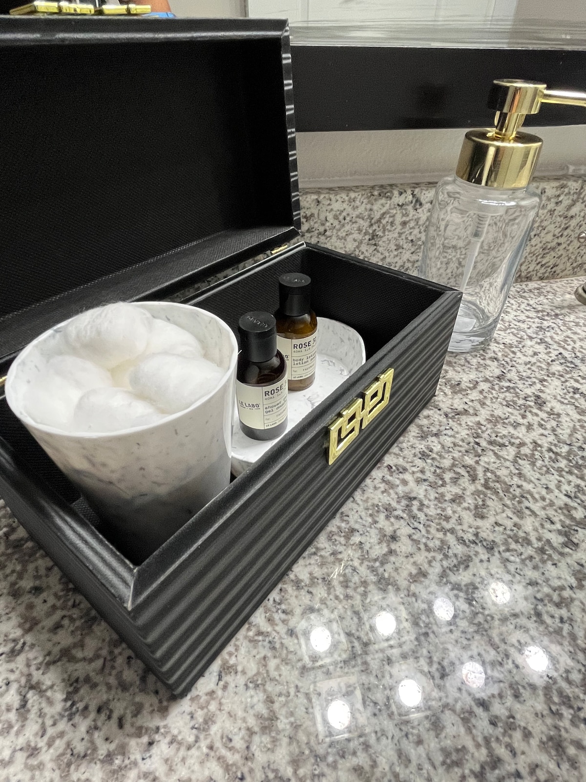 A stylish, black box is positioned on a granite countertop, containing a cup filled with white bath products. Two brown bottles labeled with essential oils are visible alongside a clear glass dispenser with a golden pump. The surroundings exhibit a sleek and modern bathroom aesthetic.