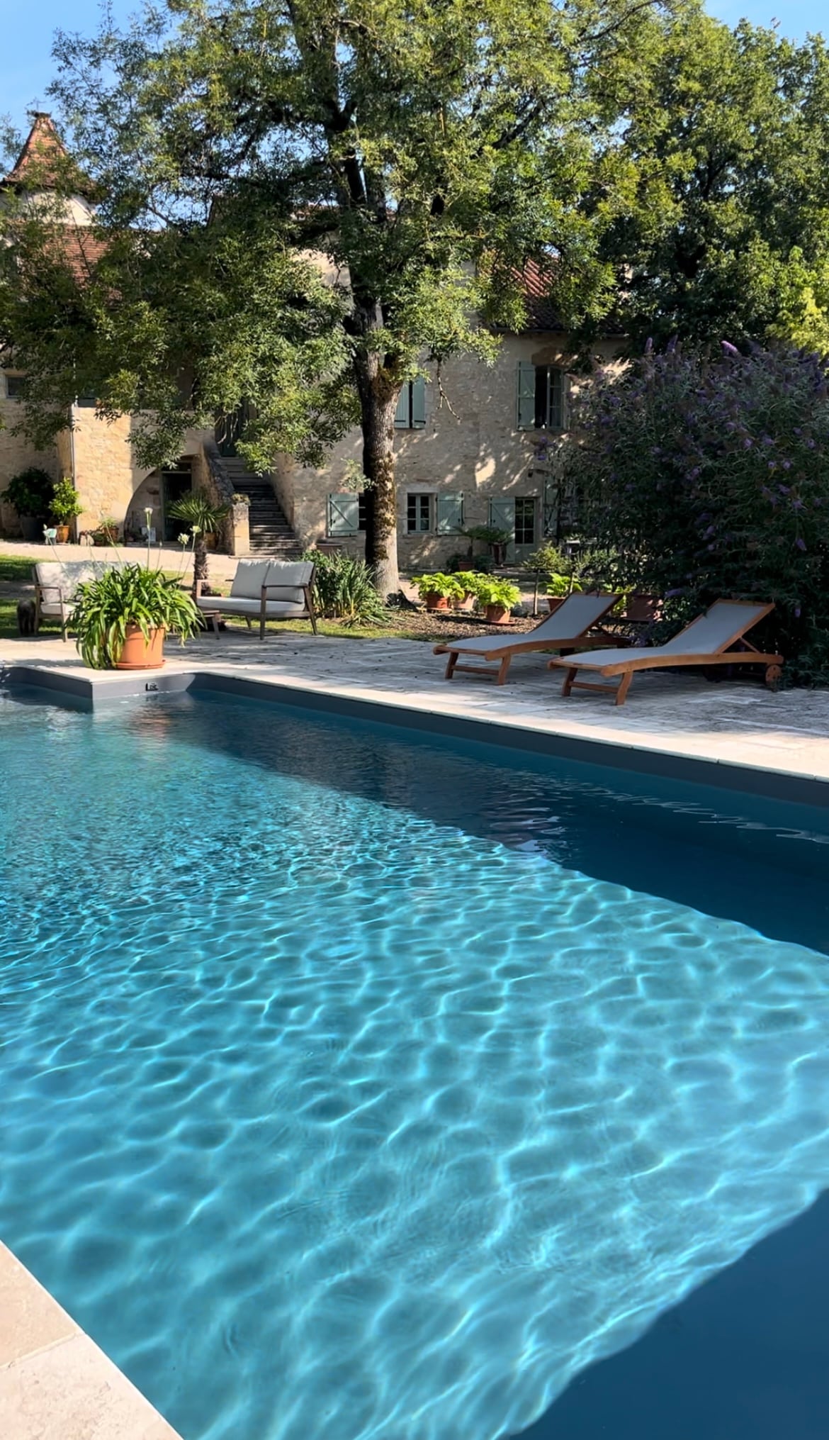 A serene outdoor area features a shimmering pool with clear blue water, surrounded by lush greenery and colorful plants. Two lounge chairs are positioned beside the pool, providing a comfortable spot for relaxation. The charming stone house is visible in the background.
