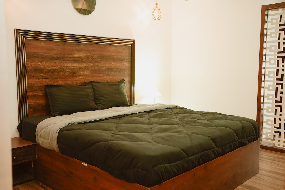 A spacious bed is centered in the room, dressed in a dark green comforter. A pair of pillows are arranged neatly against a wooden headboard. Soft lighting is provided by a bedside lamp, while a patterned screen adds a touch of texture in the background.