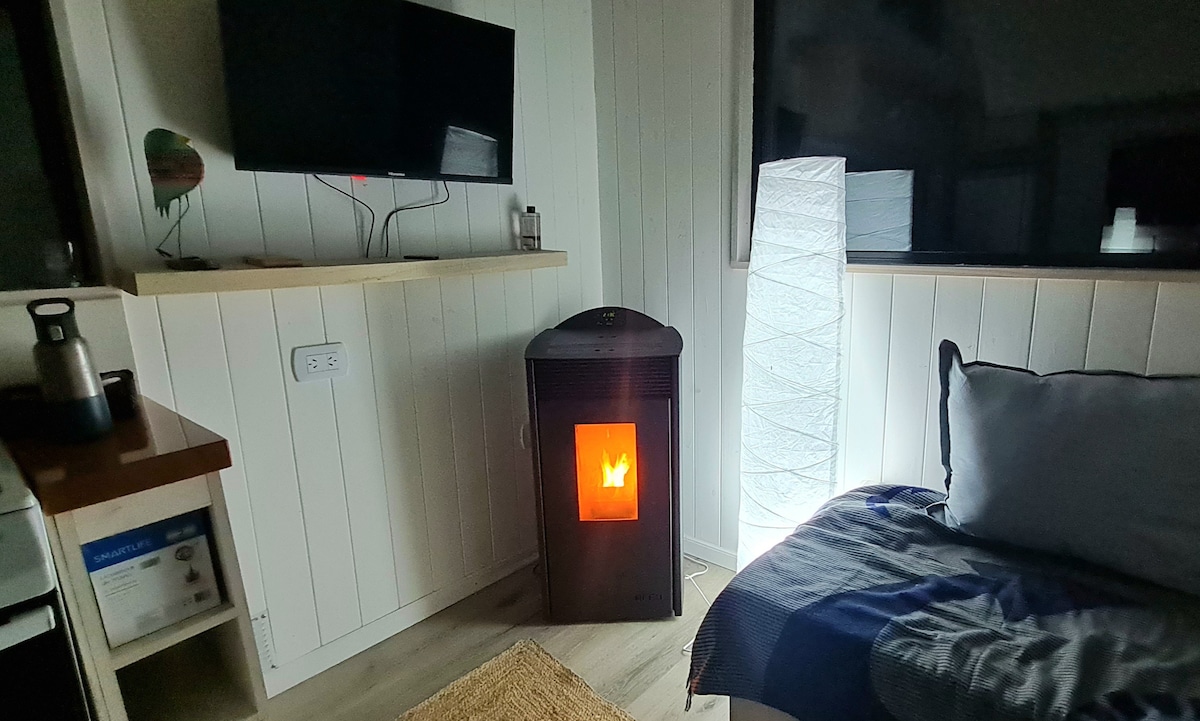 A cozy living space is highlighted by a modern electric fireplace, emitting a warm glow. A soft sofa with decorative pillows faces a large window. A textured floor rug complements the light wood flooring, while a lamp and television add functionality to the room.