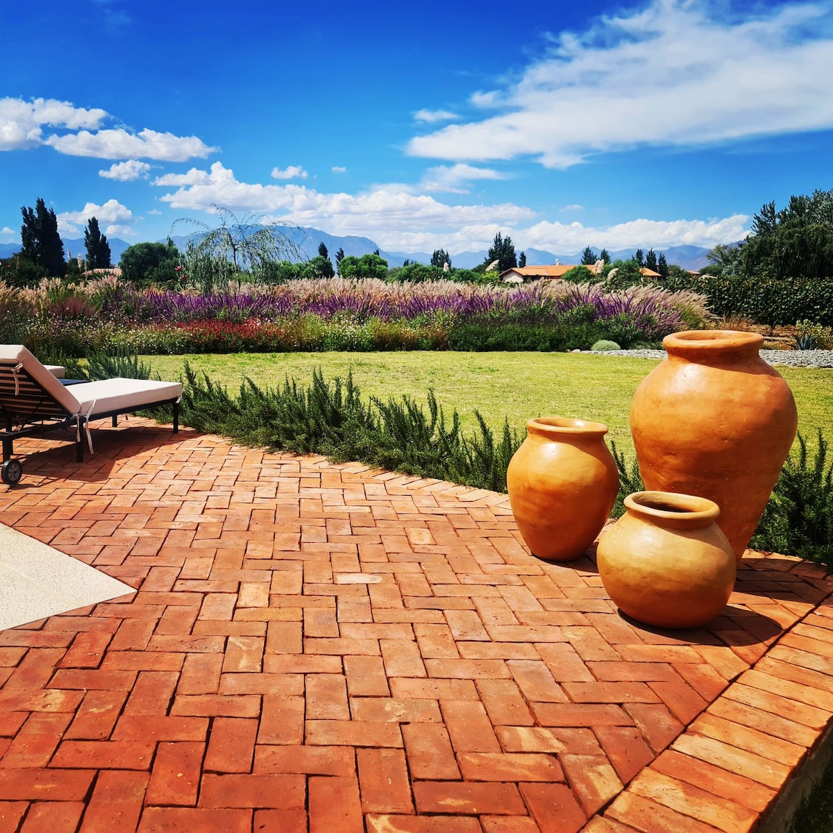 A well-tended patio features terracotta planters arranged alongside a spacious grassy area. Lush flowers in varying colors stretch across the landscape, set against a clear blue sky with scattered clouds. Sun loungers invite relaxation in the serene outdoor setting.