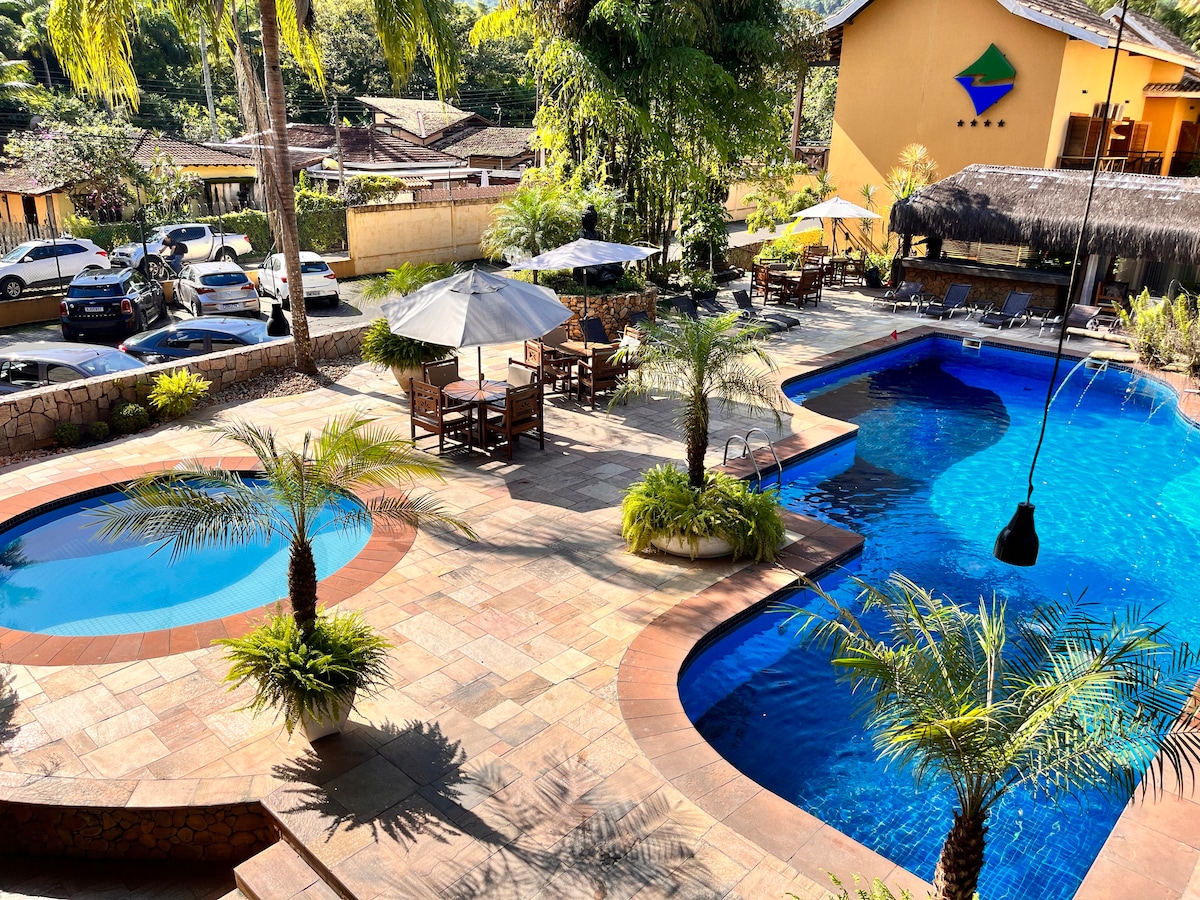 The outdoor area features two swimming pools surrounded by palm trees and lounge chairs. Umbrellas provide shade over wooden tables and chairs. The vibrant landscape is complemented by lush greenery and a well-maintained pathway, creating a serene setting for relaxation.