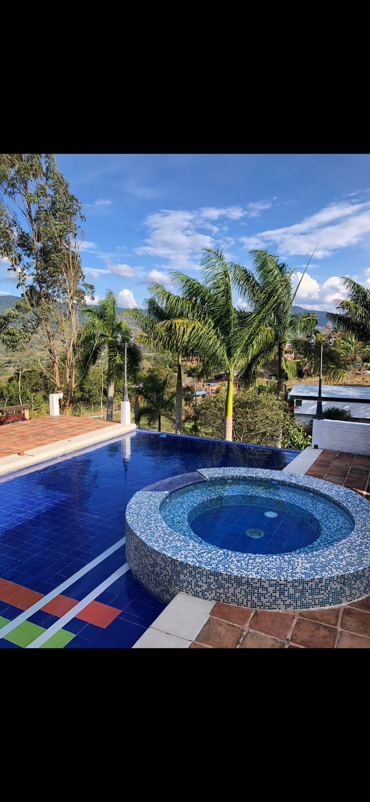 A vibrant jacuzzi is set beside a spacious pool, surrounded by lush greenery and palm trees. The clear blue water reflects the sky and mountains in the background, creating a serene outdoor space for relaxation.