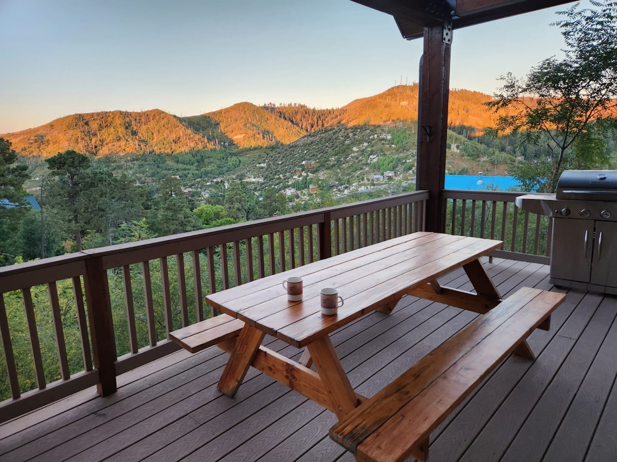A spacious wooden picnic table with two benches is situated on a deck, surrounded by beautiful mountain views. A propane gas grill is positioned nearby, and soft morning light casts a warm glow over the landscape, highlighting the serene nature of the surroundings.